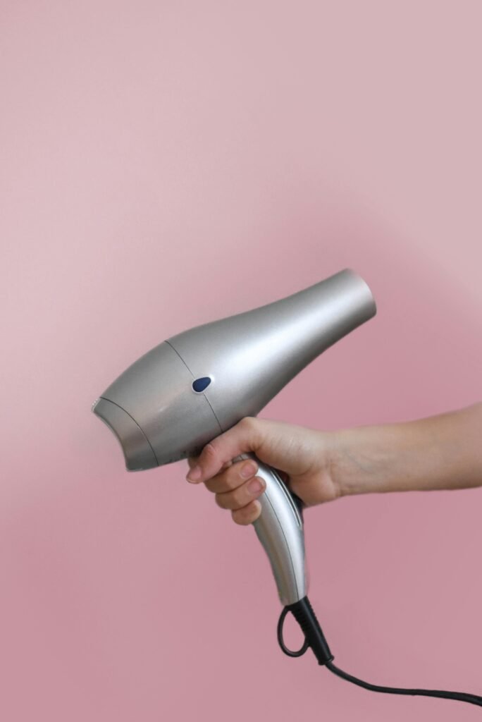 Close-up of a hand holding a silver hair dryer on a pink background.