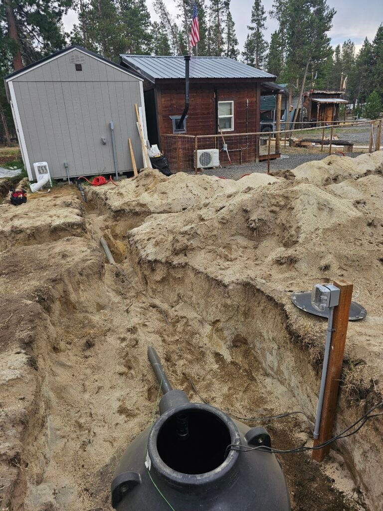 A trench dug out beside a house; inside the trench, a round septic tank is partially exposed, with a control box mounted on a wooden pole to the tank's right.